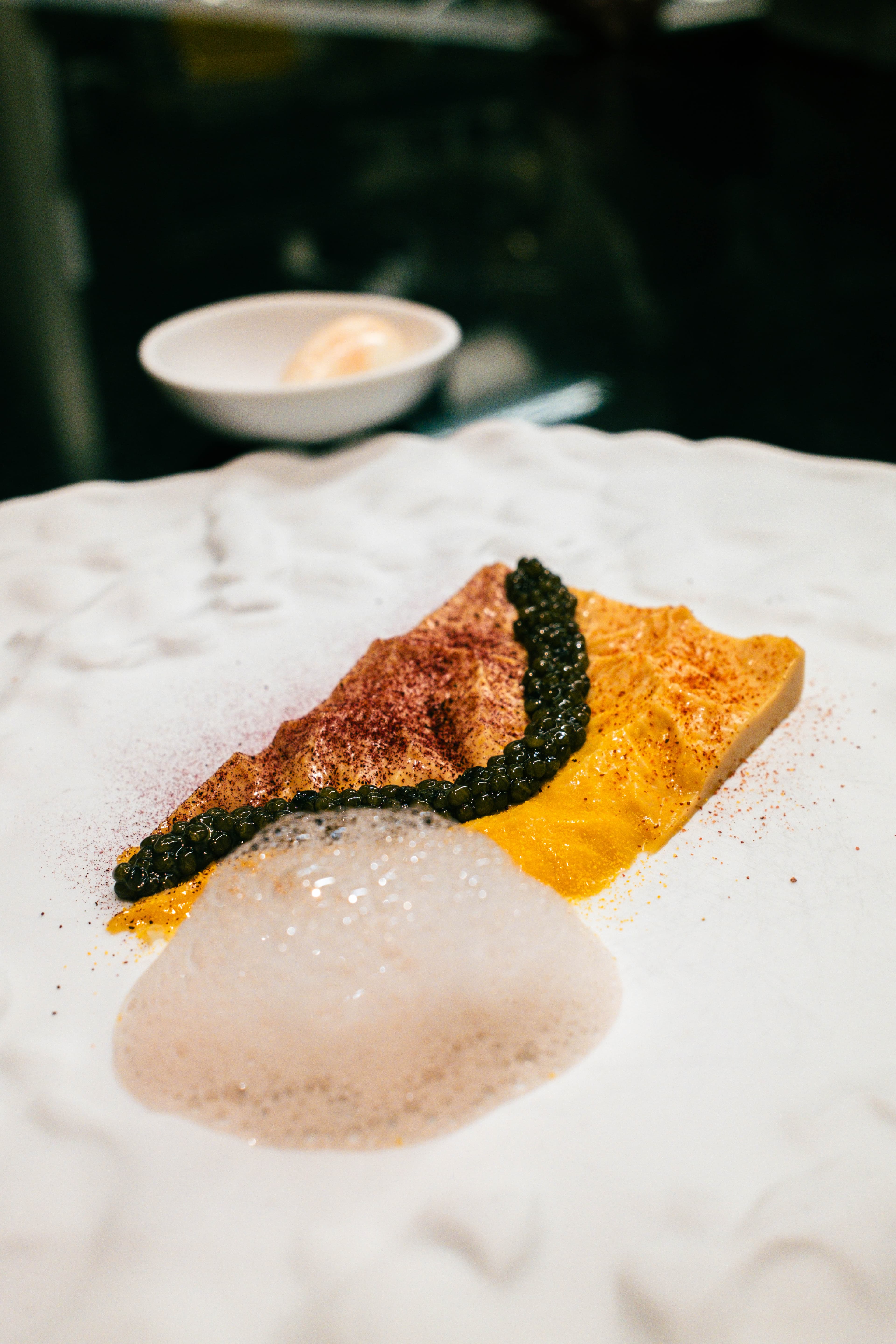  Close-up of a fine dining dish featuring caviar, a golden puree, dusted spice, and a light foam served on a white textured plate.
