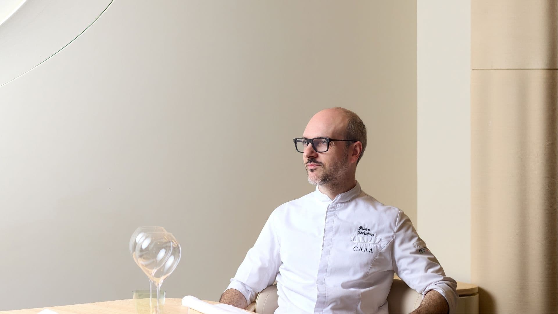 Chef Pietro Catalano in a white chef's jacket, looking thoughtfully to the side while seated in the minimalist, neutral-toned dining room of his restaurant, CAAA.