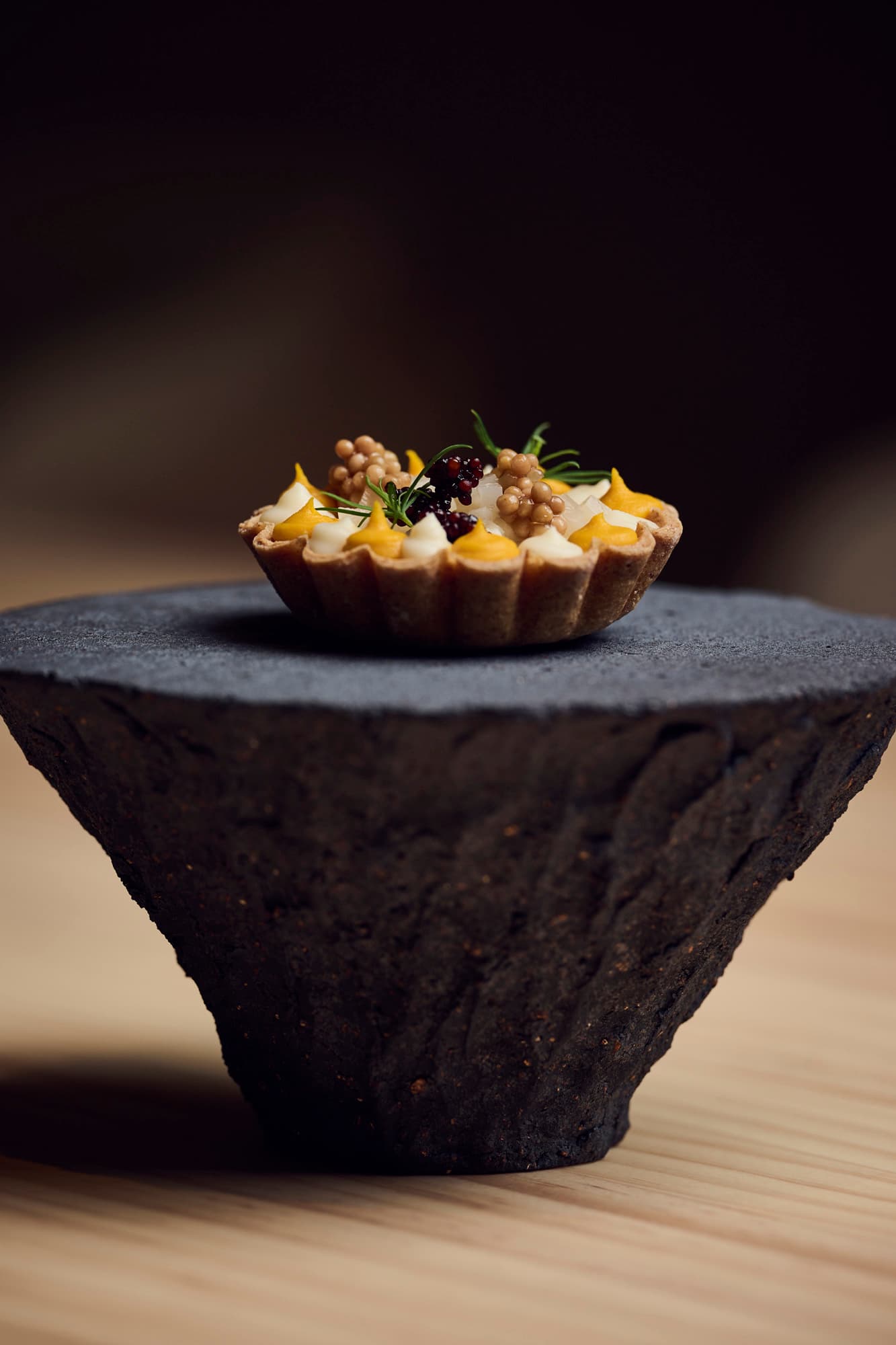 A delicate, savory tartlet filled with yellow and white dollops of cream, topped with caviar, pearls, and fresh herbs, served on a textured, dark stone pedestal.