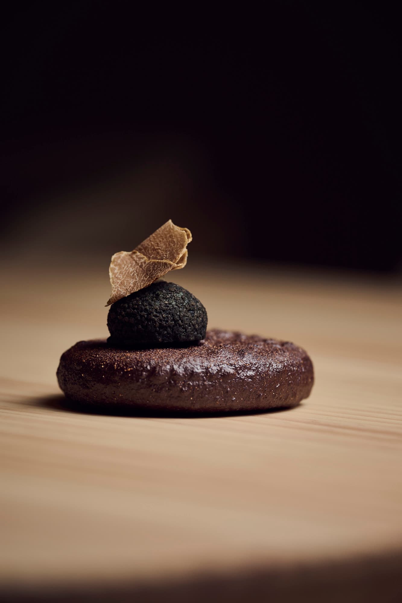 A dark round pastry base topped with a textured sphere and a thin dried garnish, photographed on a warm wood surface with a dark backdrop