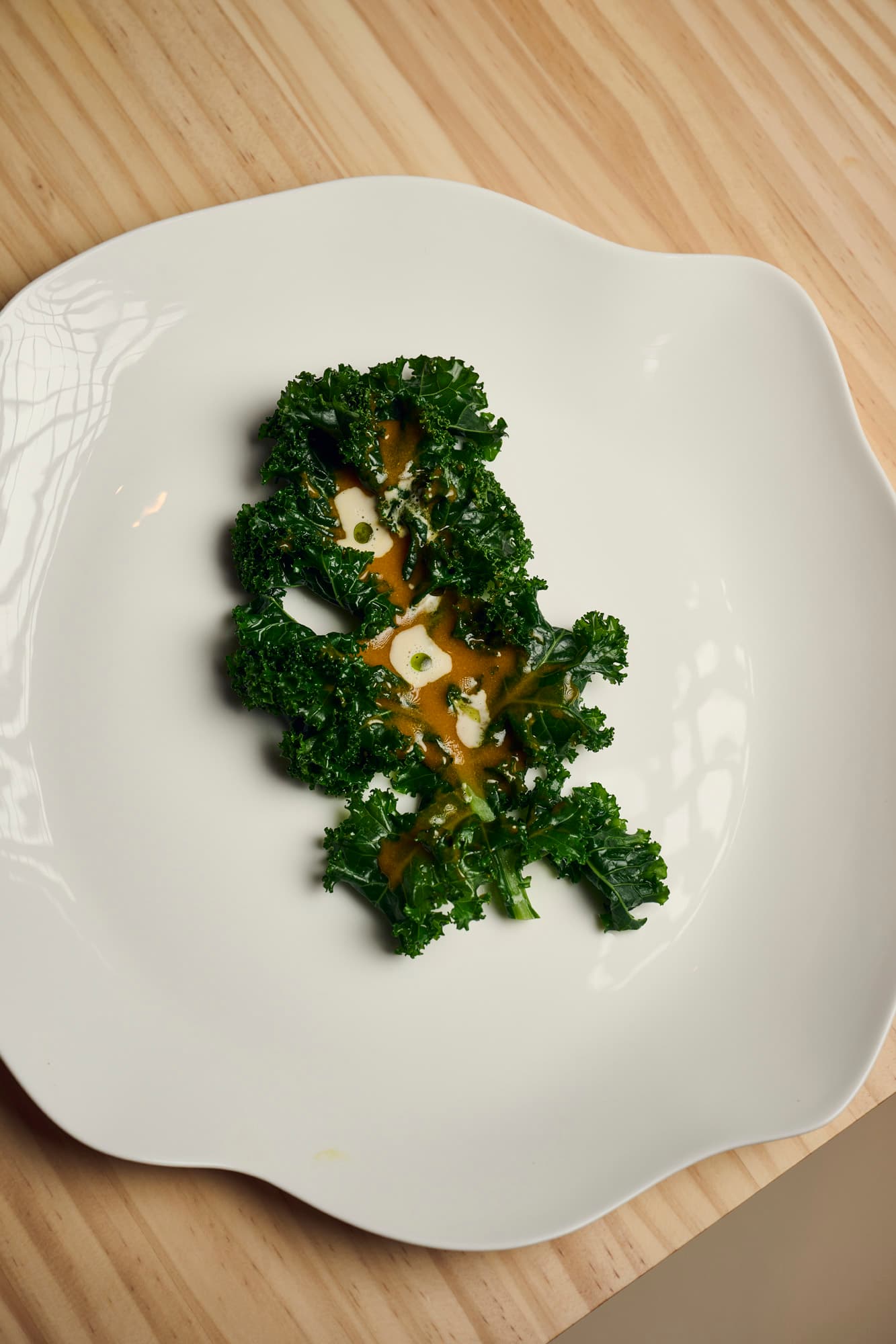 Overhead view of a plated kale dish with sauce on a white sculptural plate set on a light wood surface at a fine dining restaurant.
