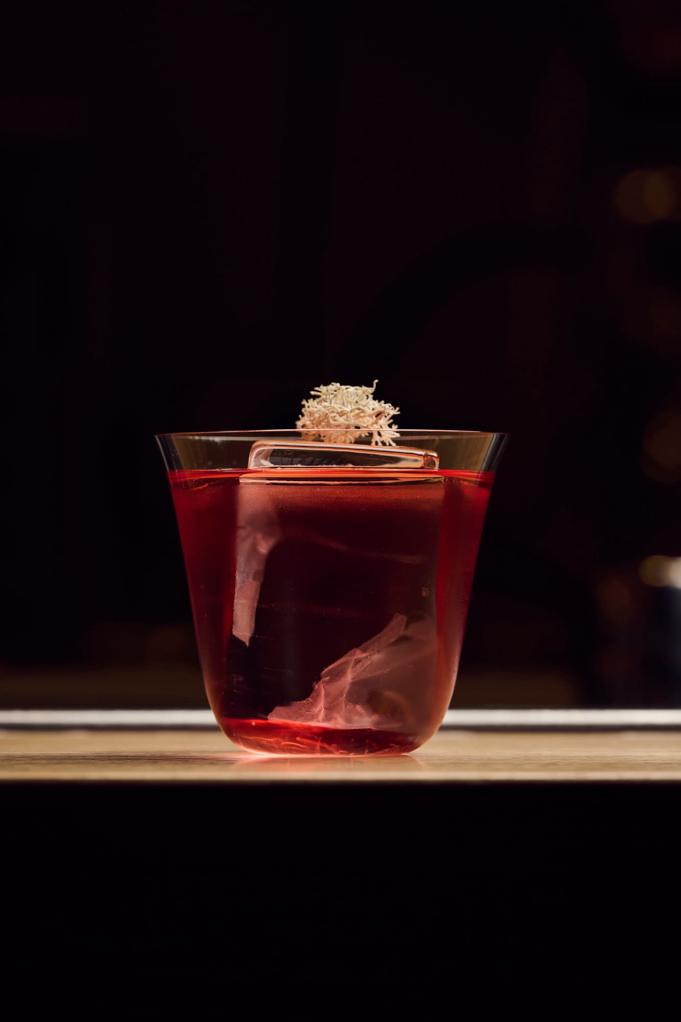 A deep red cocktail served over a large ice cube in a clear glass, garnished with a dried botanical, set against a dark background.