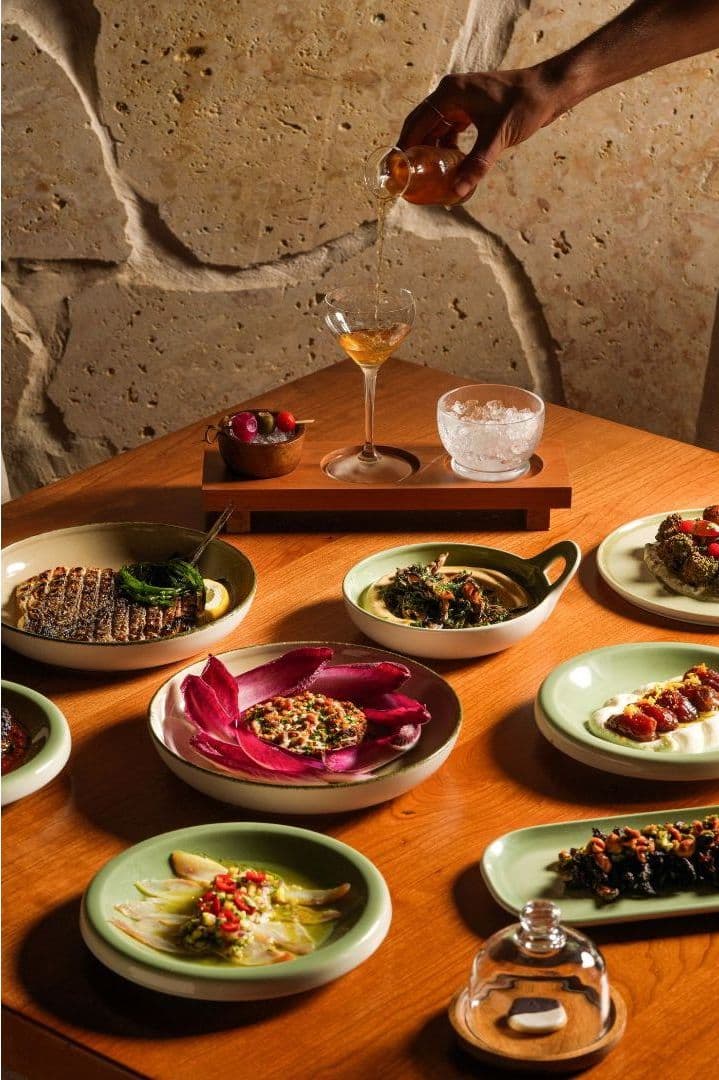 Colorful spread of Mediterranean dishes on a wooden table with a hand pouring a golden drink into a coupe glass against a travertine stone wall