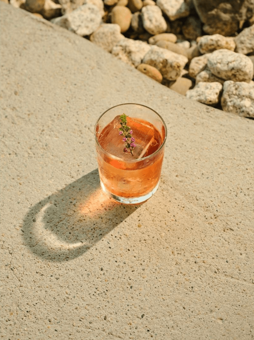 Amber-hued cocktail with a large ice cube and fresh herb garnish resting on a sunlit stone ledge