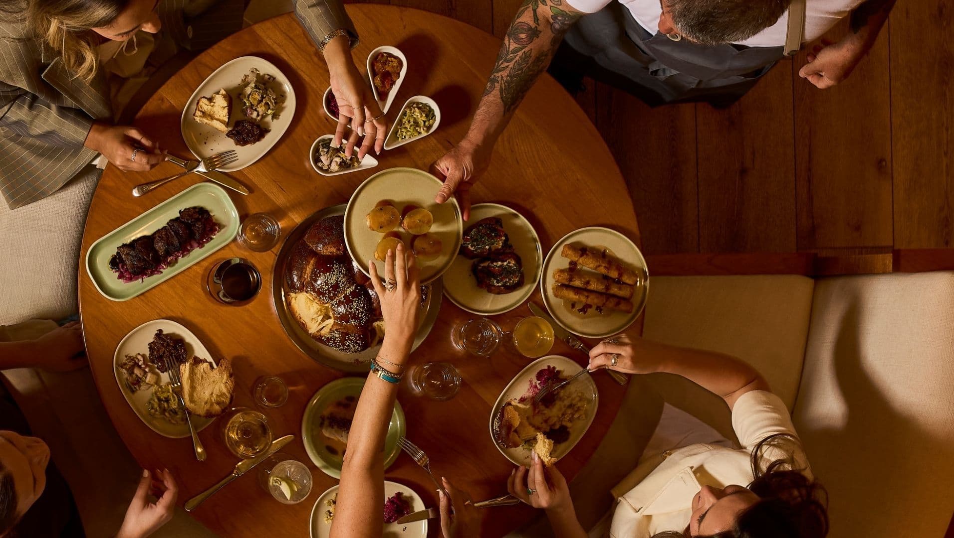 Overhead view of guests sharing plates of food family-style at a round wooden table in a warmly lit restaurant