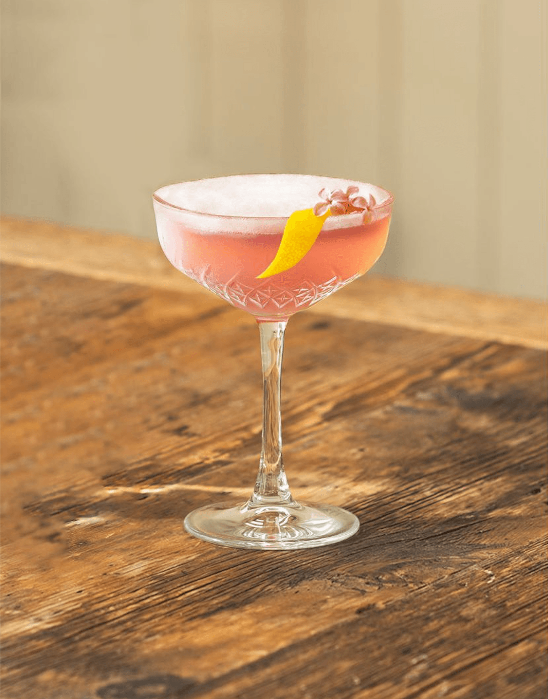 Pink cocktail in a crystal coupe glass garnished with a lemon peel twist and small flower on a rustic wooden table