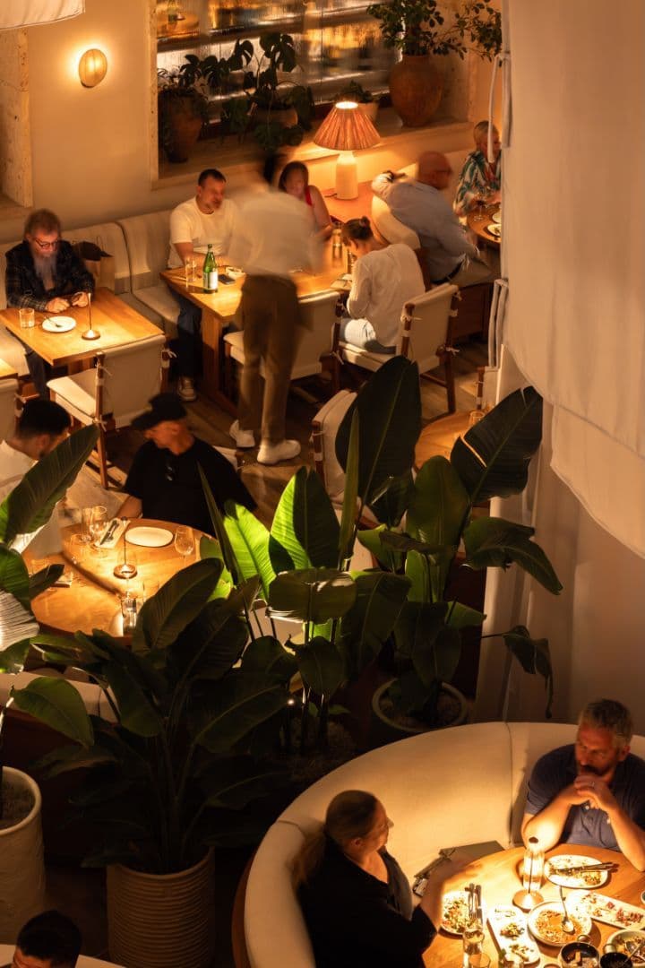 Overhead view of a warmly lit hotel restaurant at night with guests dining at intimate tables among tropical plants and draped curtains