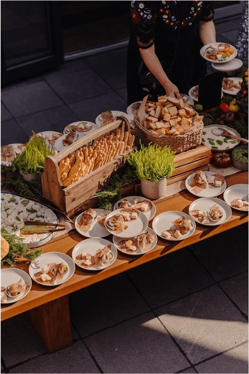 Outdoor event buffet table with individually plated charcuterie, artisan breadsticks, fresh mozzarella, and seasonal vegetables on a wooden display