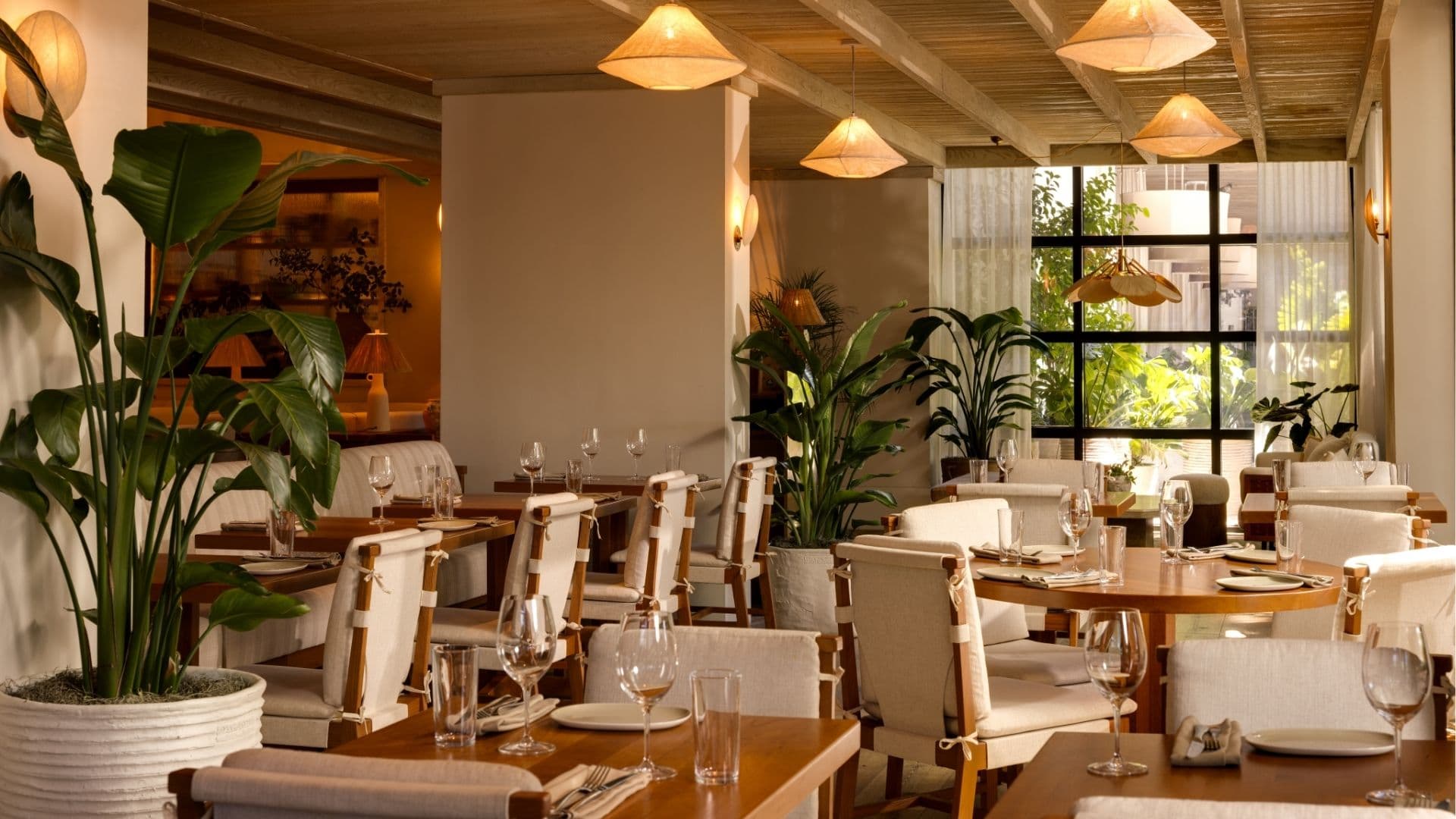Sunlit hotel restaurant dining room with natural wood furniture, linen-wrapped chairs, cone-shaped pendant lights, and tropical plants